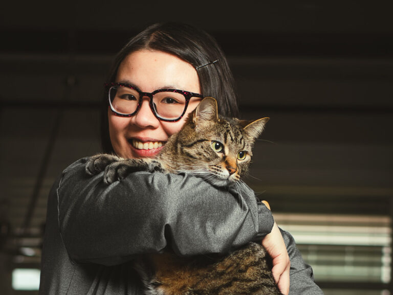 A woman wearing glasses looks at the camera while holding a tabby cat